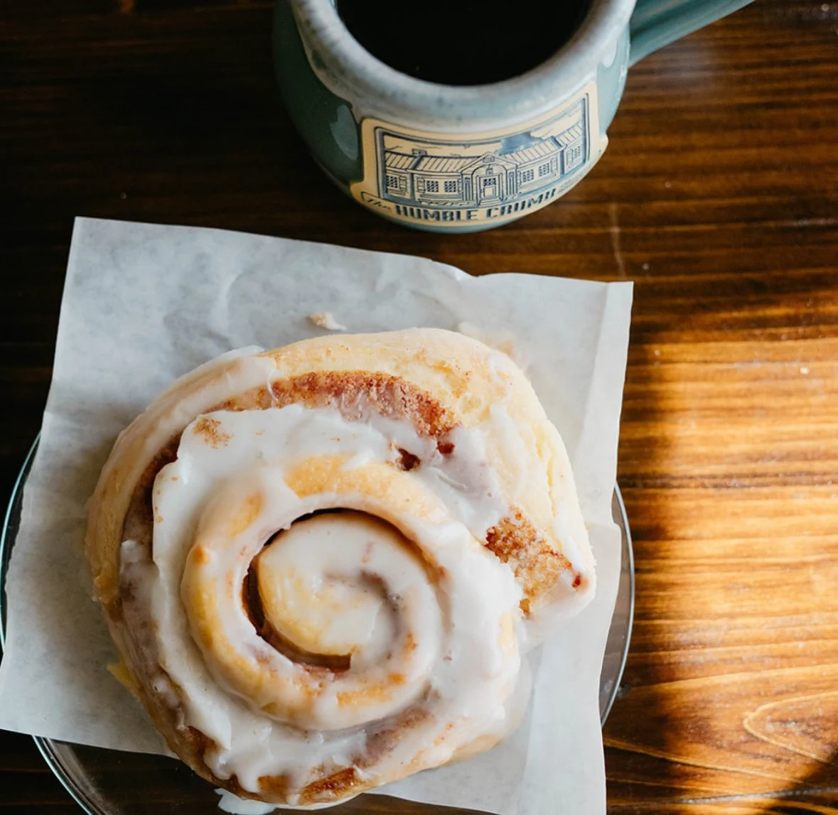 An iced Humble Crumb cinnamon roll on parchment beside a bakery mug